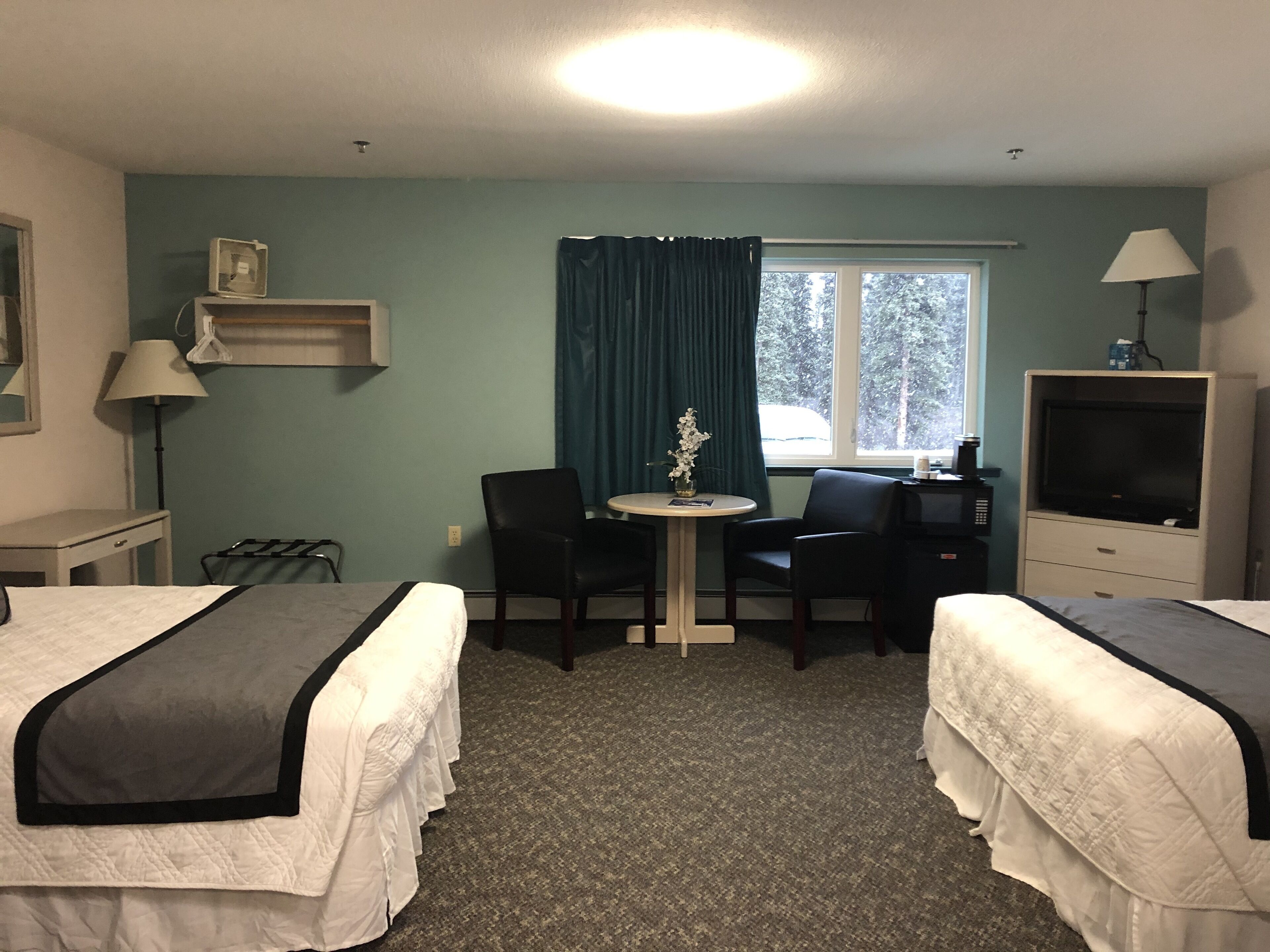 A hotel room with two beds, a desk, a chair, a small table with a vase, a TV, and a window with curtains.