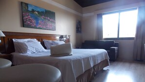 A hotel room with a bed, a desk, a chair, a window, and a painting on the wall. - Raupp Flats Gramado 01 (Gramado)