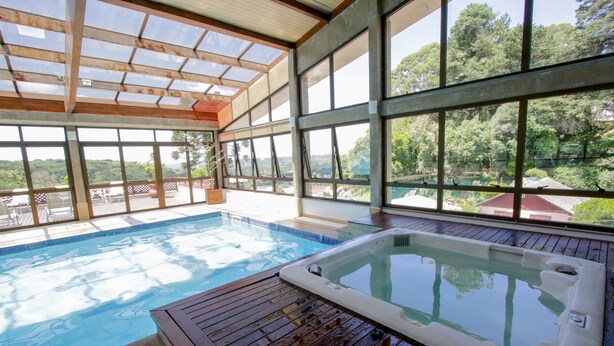 A swimming pool with a hot tub, wooden deck, and large windows offering a view of the surrounding landscape. - Raupp Flats Gramado 01 (Gramado)