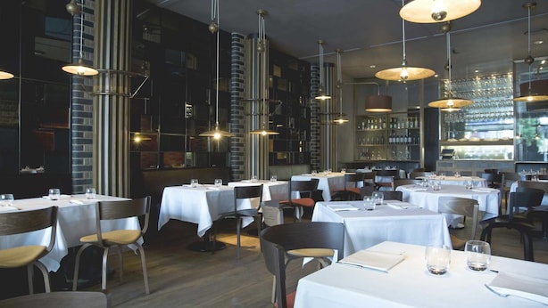 A restaurant interior with multiple tables, chairs, hanging pendant lights, and a bar area with shelves of bottles.