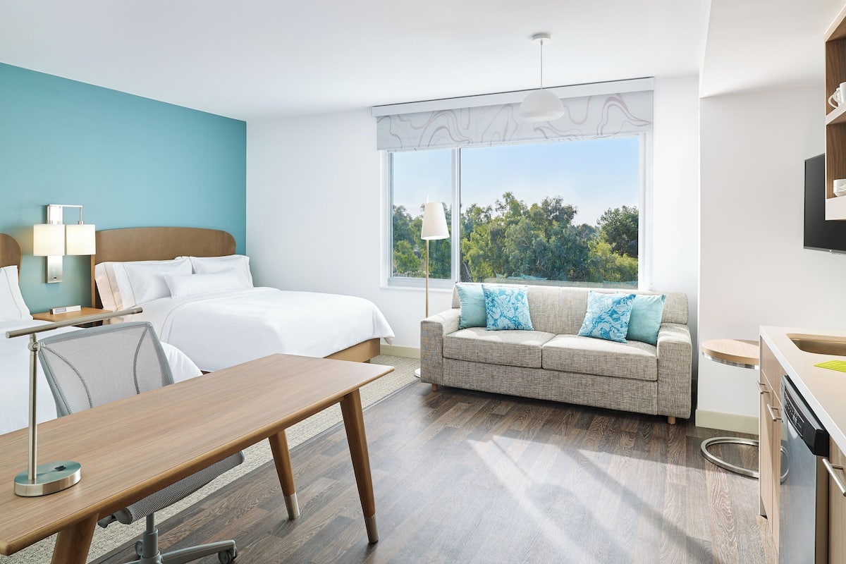 Een hotelkamer met een bed, een bank, een bureau, een lamp en een raam met uitzicht op bomen.