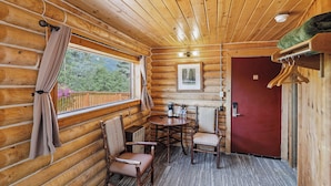 A wooden cabin interior with a dining table, two chairs, a red door, and a window with curtains.