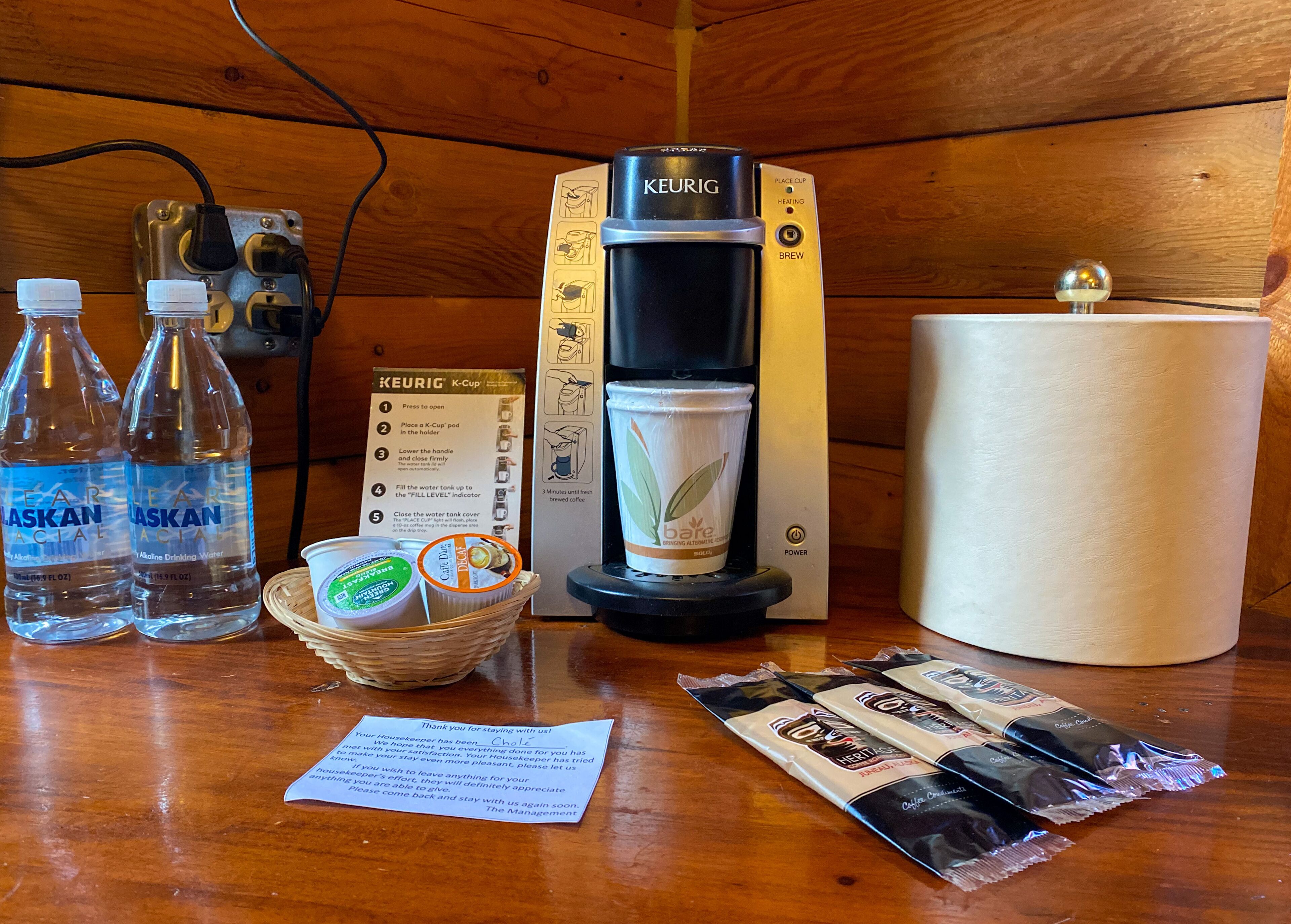 A coffee maker, water bottles, a cup of coffee, a basket with snacks, and a note on a wooden table.