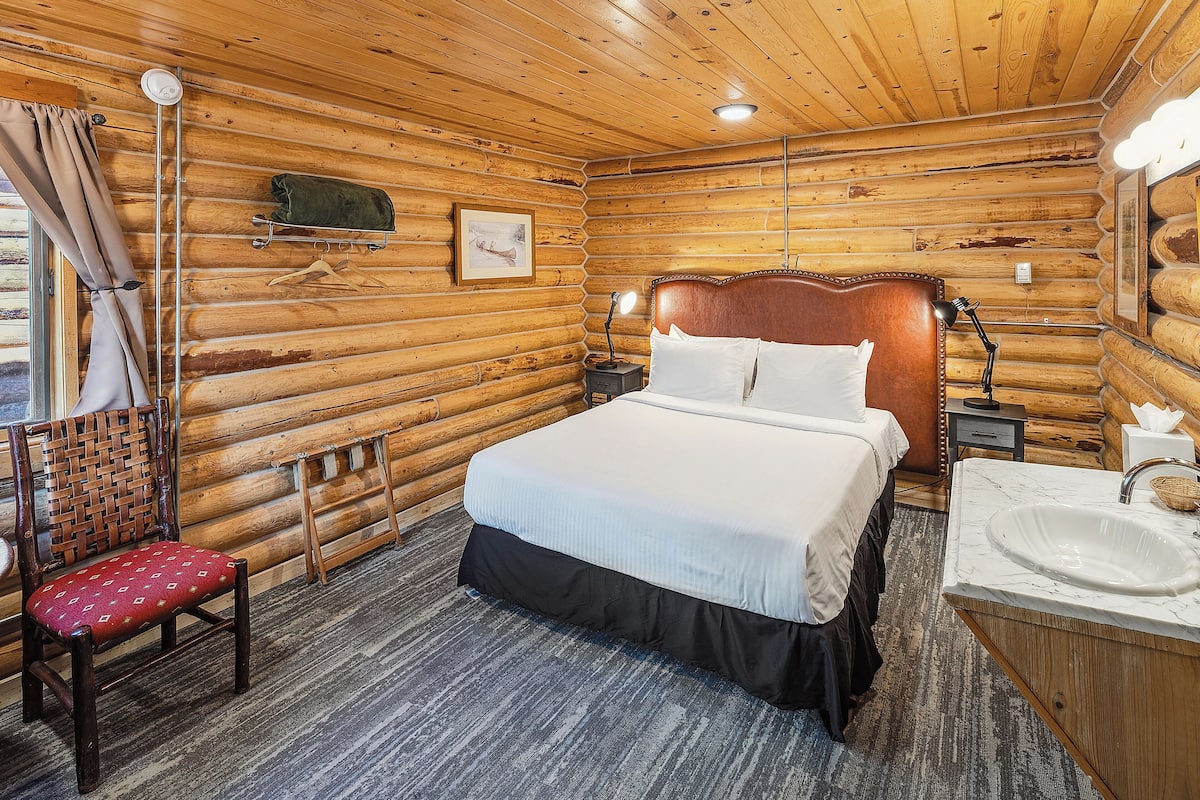 A log cabin bedroom with a bed, bedside table, lamp, chair, and a bathroom with a sink.