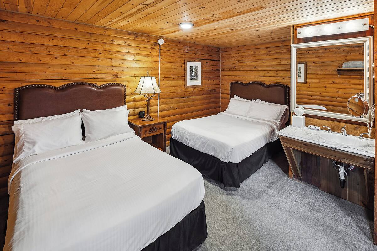 A wooden-paneled room with two beds, a nightstand, a lamp, a mirror, and a sink.