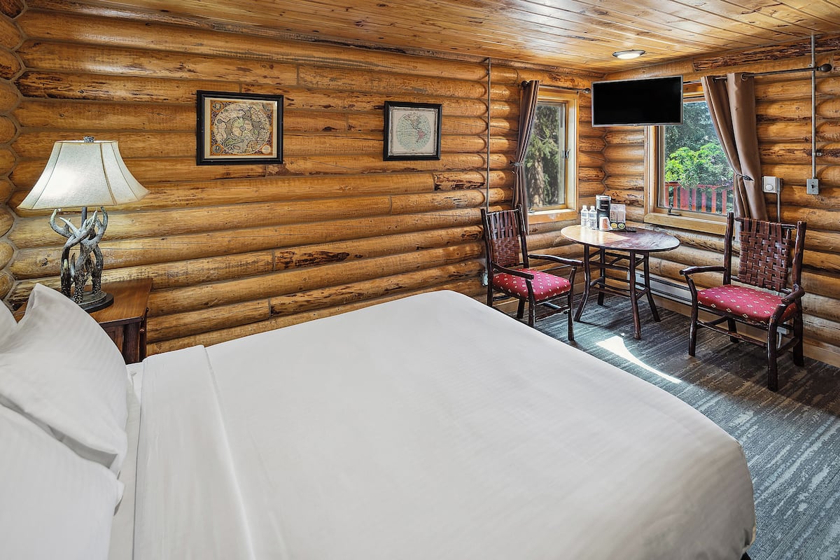A log cabin room with a bed, a table with chairs, a lamp, and two framed pictures on the wall.