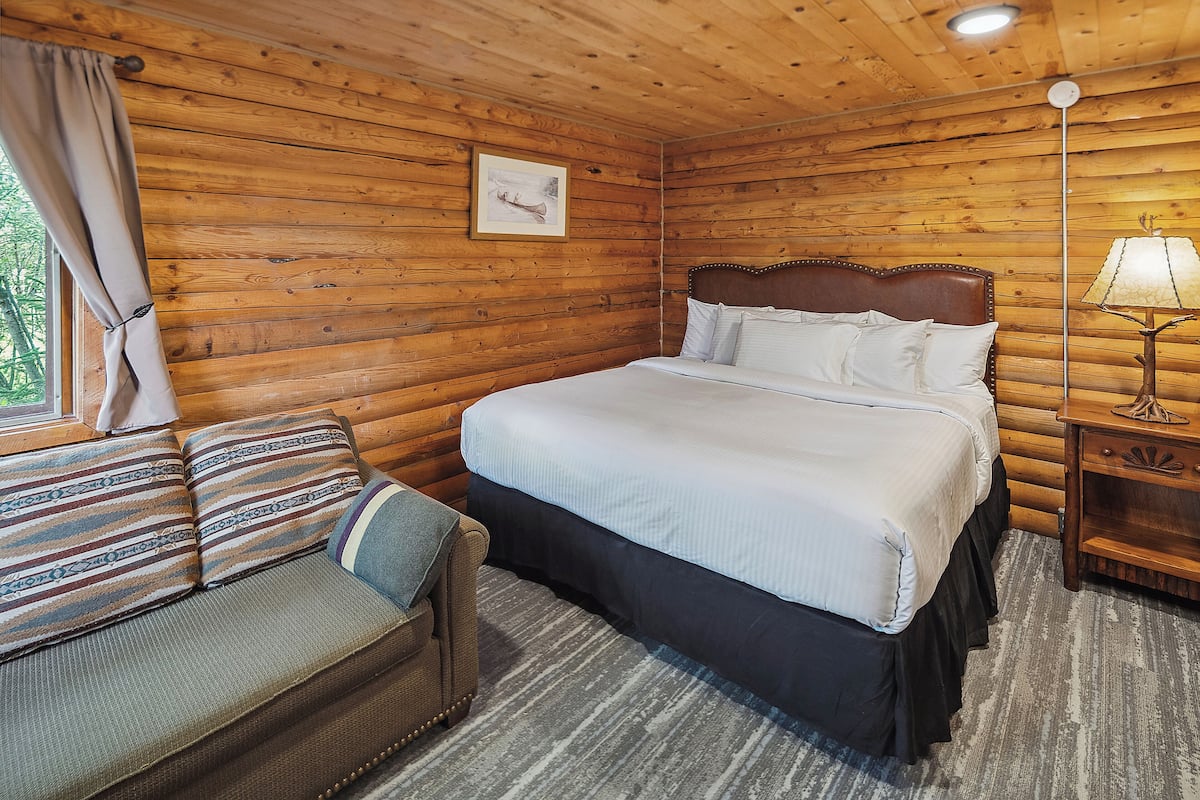 A wooden cabin bedroom with a bed, a sofa, a lamp, and a picture on the wall.
