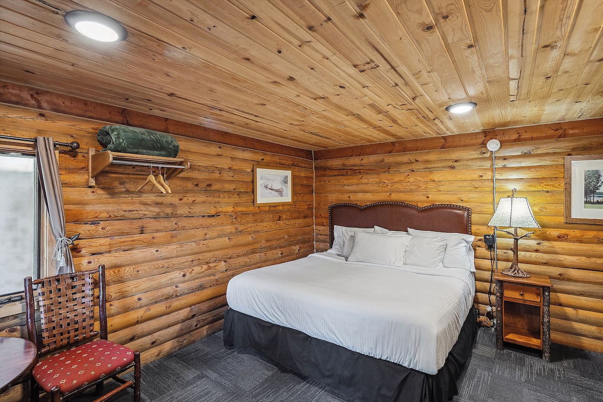 A wooden cabin room with a bed, a chair, a table, and a lamp.