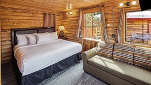 A log cabin room with a bed, a sofa, a window with curtains, and a table with chairs outside.