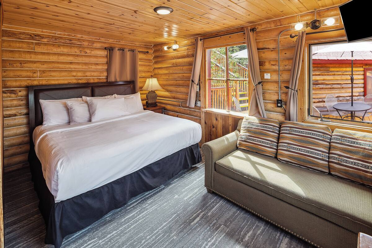 A log cabin room with a bed, a sofa, a window with curtains, and a table with chairs outside.