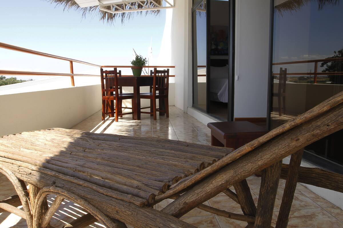 Una silla de lounge de madera en un balcón con una mesa y sillas, una planta en maceta y vistas del entorno.