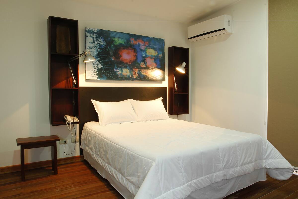 Una cama bien hecha con ropa blanca, una mesita de noche, un teléfono en la pared y un cuadro abstracto colorido sobre la cama.