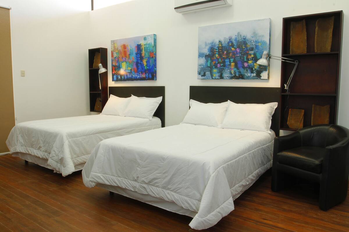 Una habitación de hotel con dos camas, un sillón de cuero negro y dos cuadros abstractos en la pared.