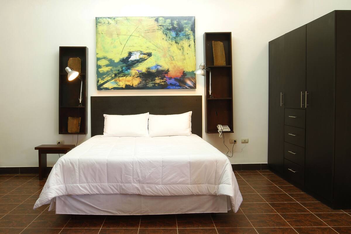 Una cama bien hecha con ropa de cama blanca, un cuadro abstracto grande en la pared, una mesita de noche con una lámpara y un armario con varios cajones.