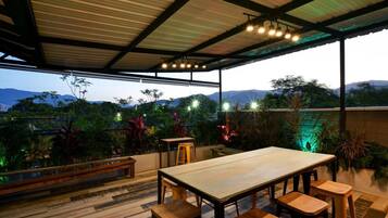 An outdoor dining area with a wooden table and chairs, surrounded by lush greenery and a view of distant mountains.