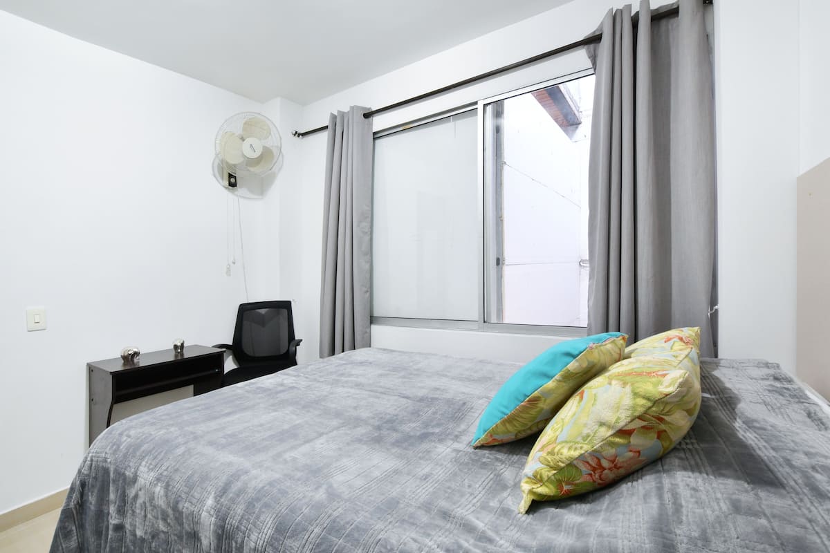 Un dormitorio con cama, escritorio, silla, ventilador y ventana con cortinas.