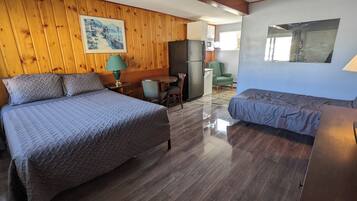 Una habitación con una cama, una cama adicional, una mesa con sillas, un refrigerador y un cuadro en la pared.