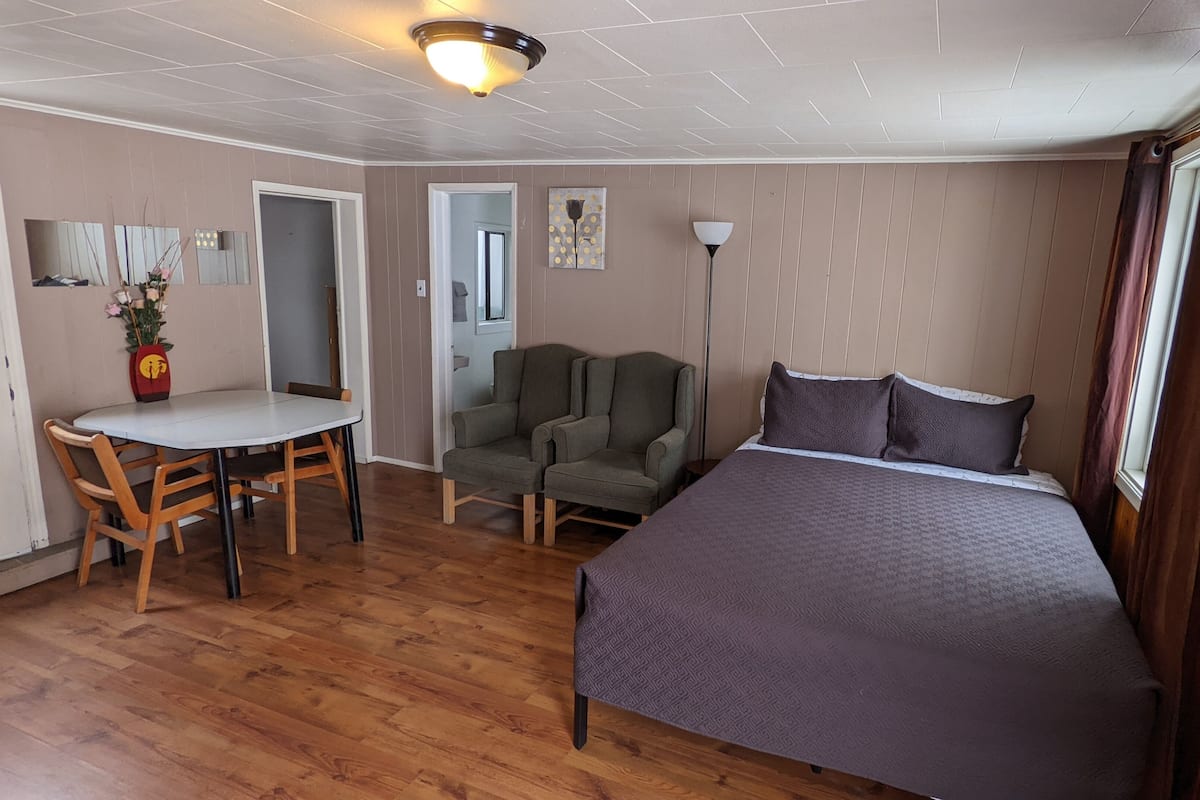 A room with a bed, a desk with chairs, a lamp, and a door.