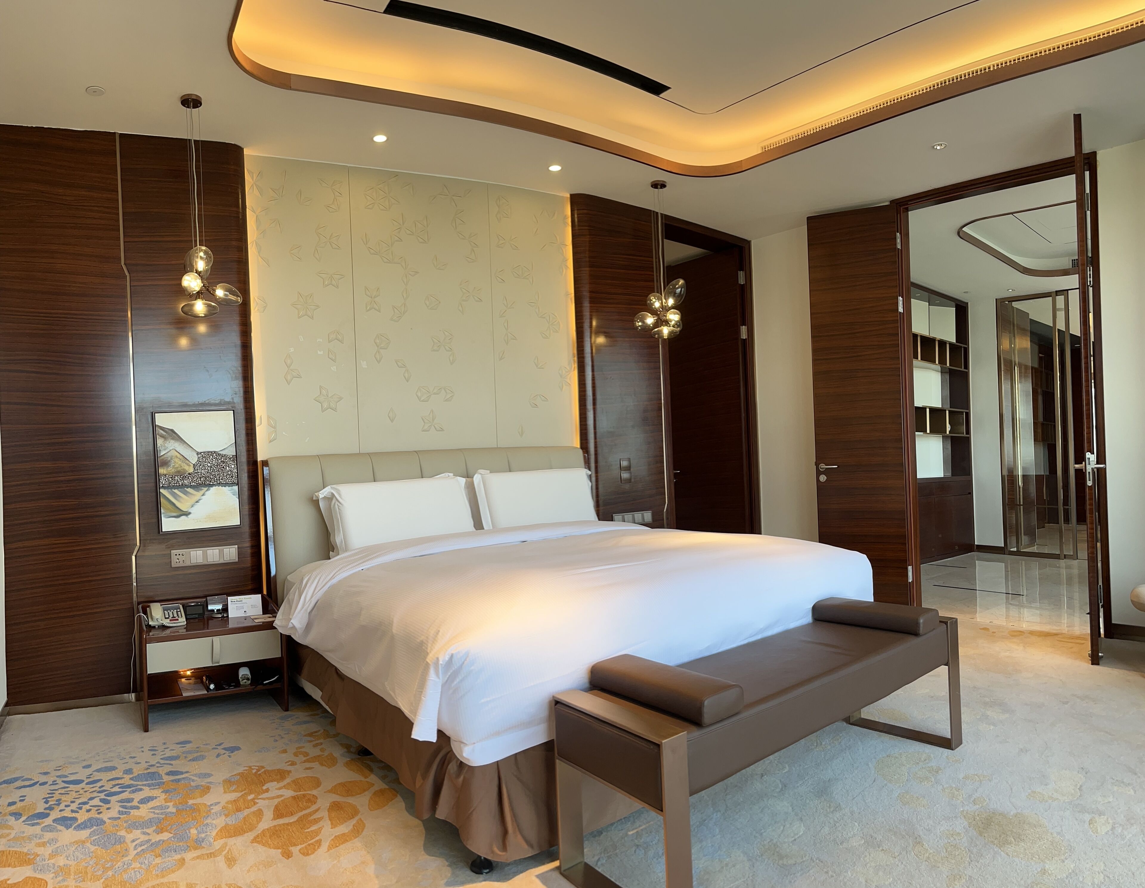 A modern bedroom with a large bed, bedside tables, a bench, a wardrobe, and a mirror.
