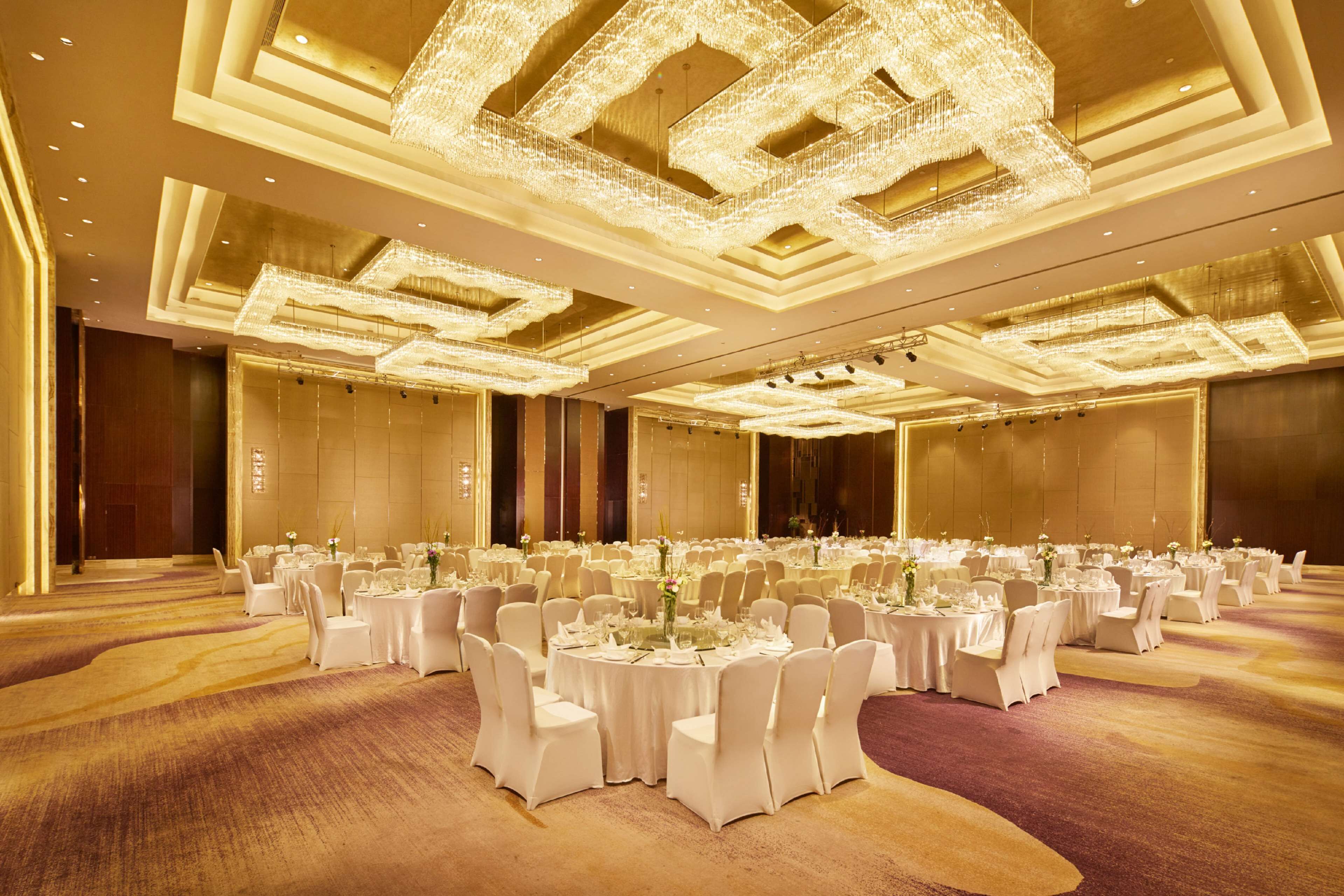 Uma sala de banquetes com mesas redondas, cadeiras brancas e lustres.