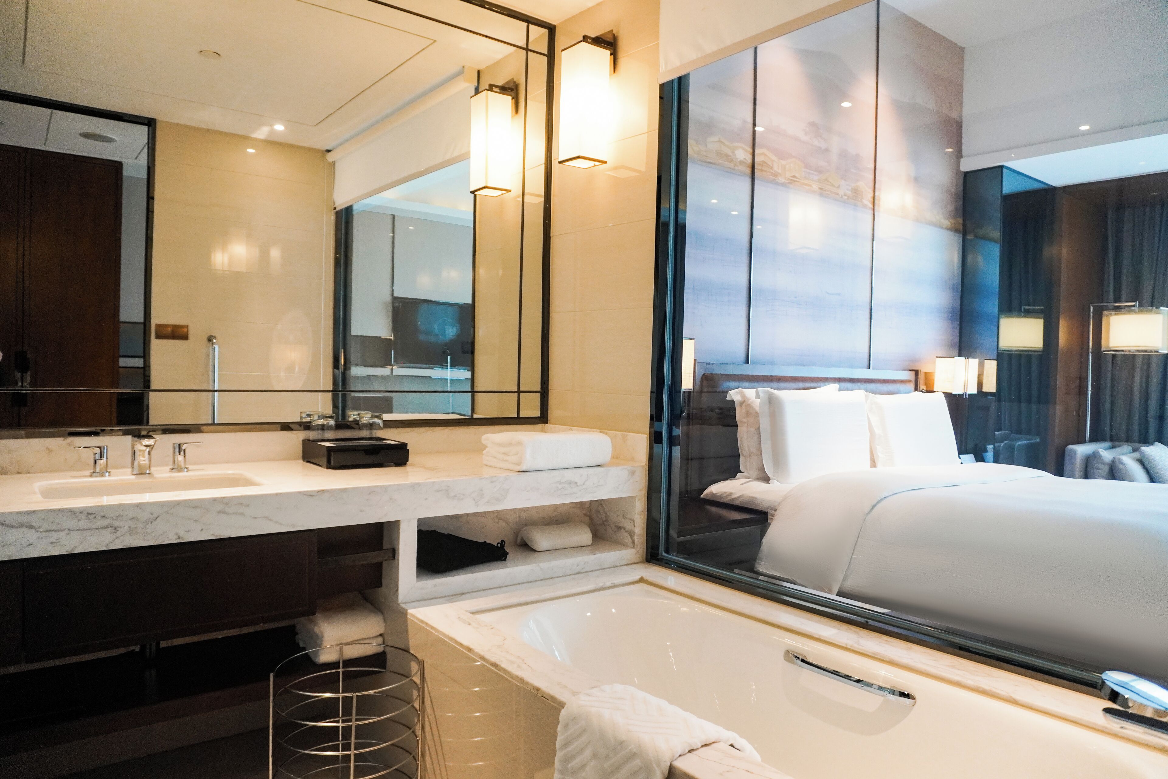 A modern hotel bathroom with a large mirror, two sinks, a bathtub, and a large bed with a view of the city.