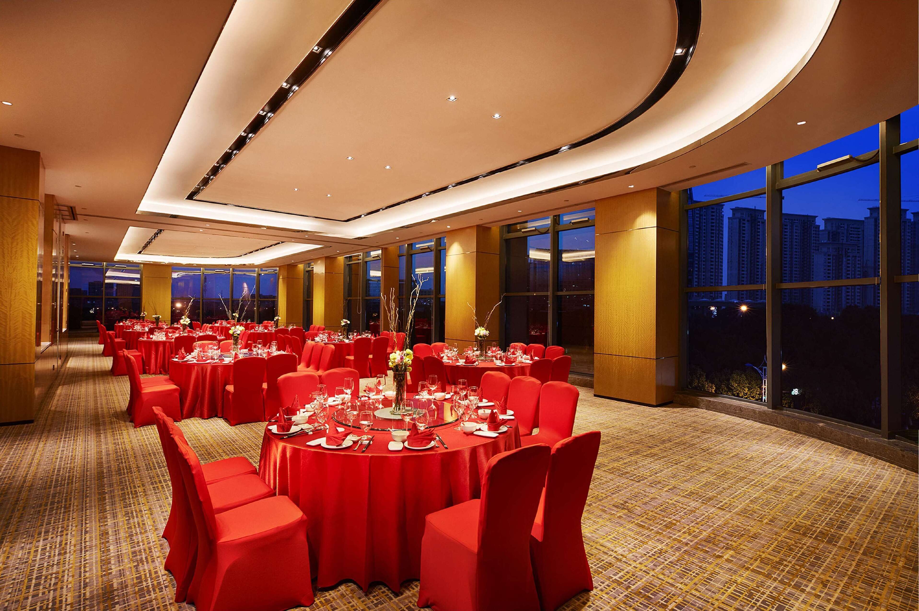 A spacious banquet hall with round tables, red tablecloths, and chairs, featuring a high ceiling and large windows with a city view.