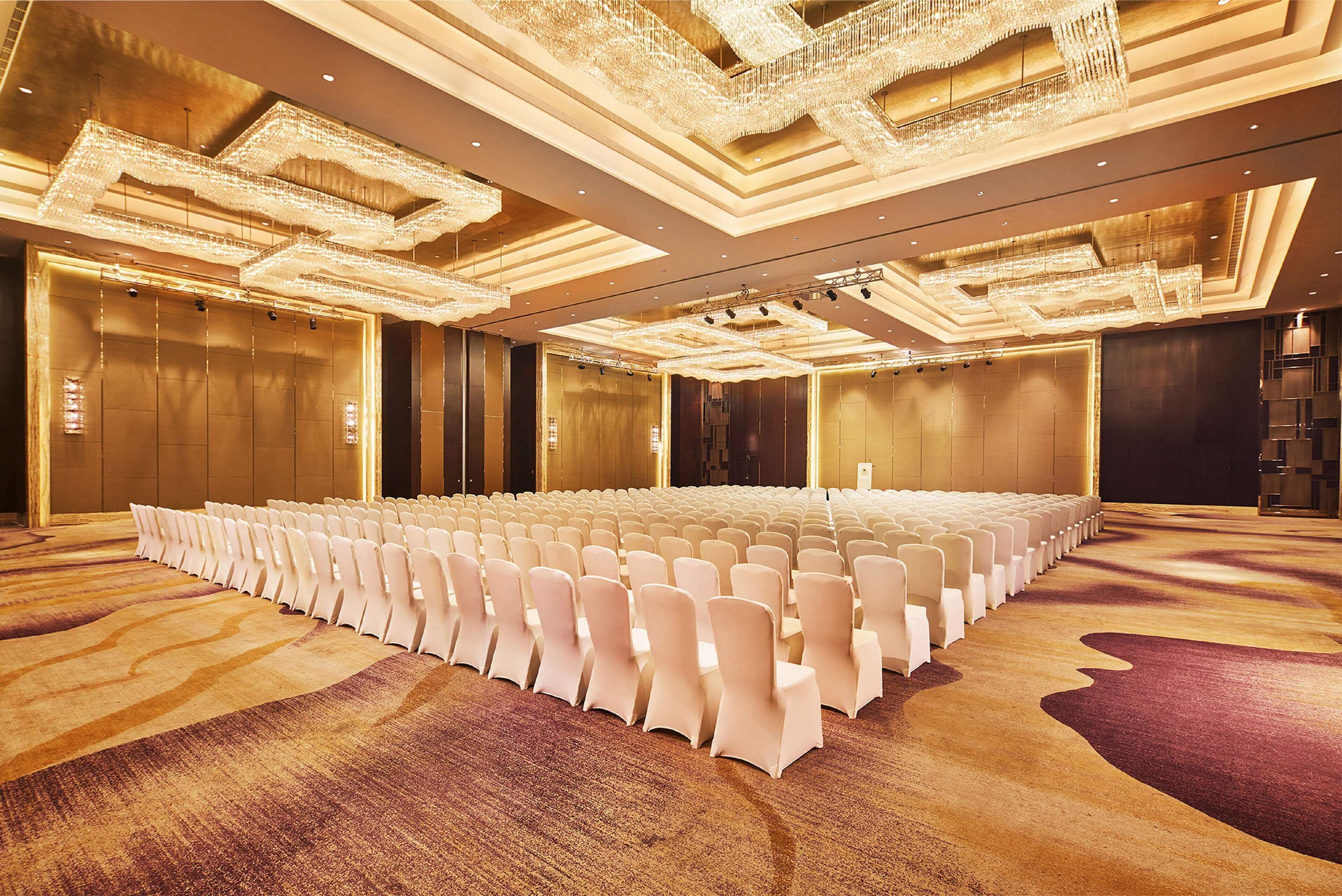 A large conference room with rows of white chairs, a patterned carpet, and ornate chandeliers.