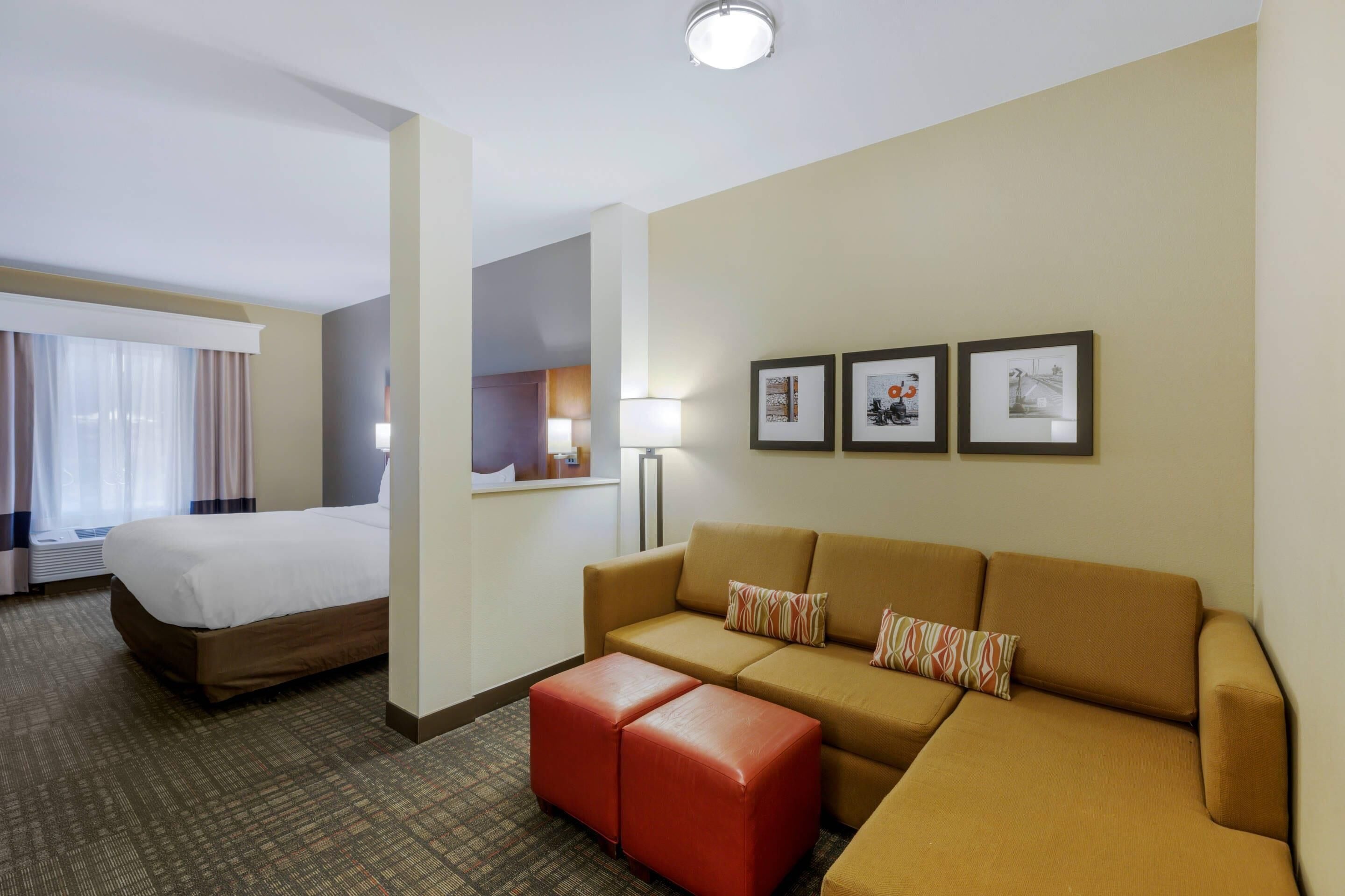 A hotel room with a bed, a sofa, a red ottoman, a lamp, and three framed pictures on the wall.
