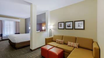 Quarto de hotel com cama, sofá, um banquinho vermelho, uma luminária e três quadros na parede.