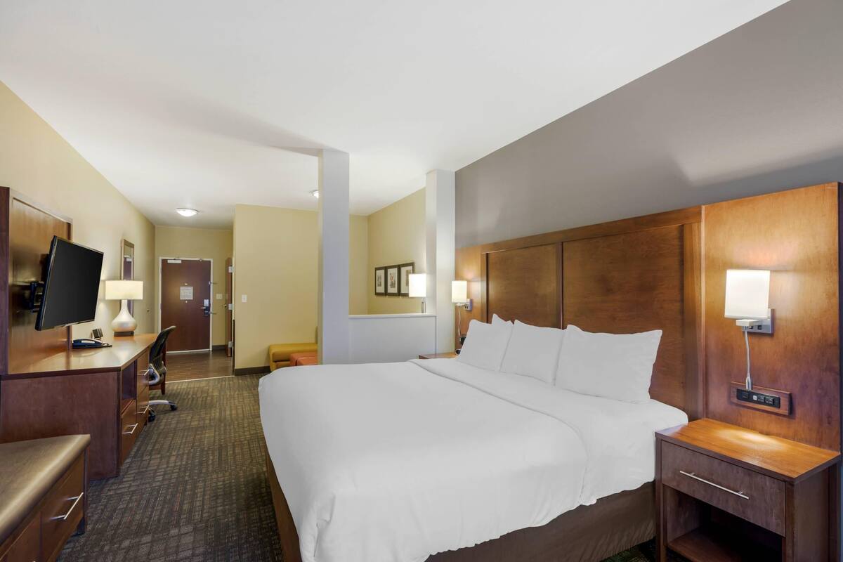 A hotel room with a large bed, a desk with a computer, a chair, a TV, and a bedside table with a lamp.