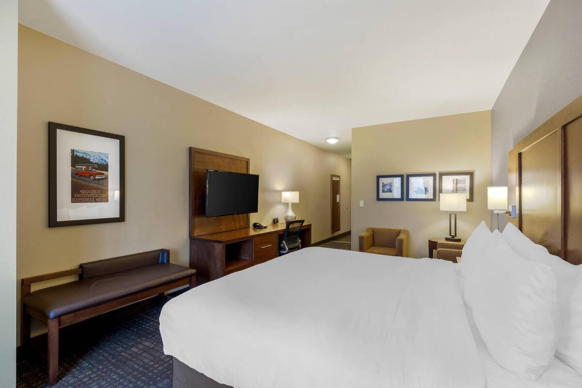 A hotel room with a large bed, a desk with a flat-screen TV, a bench, and a wall-mounted TV.