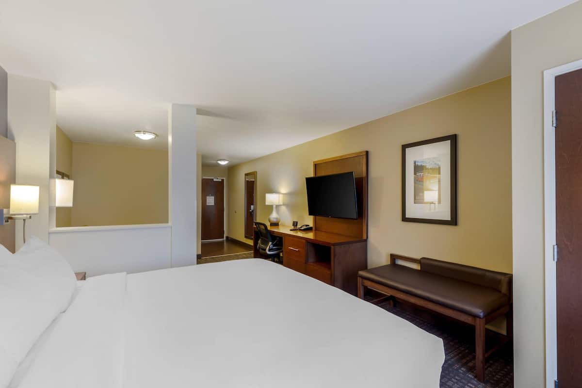 A hotel room with a large bed, a desk with a chair, a television, a bench, and a door.