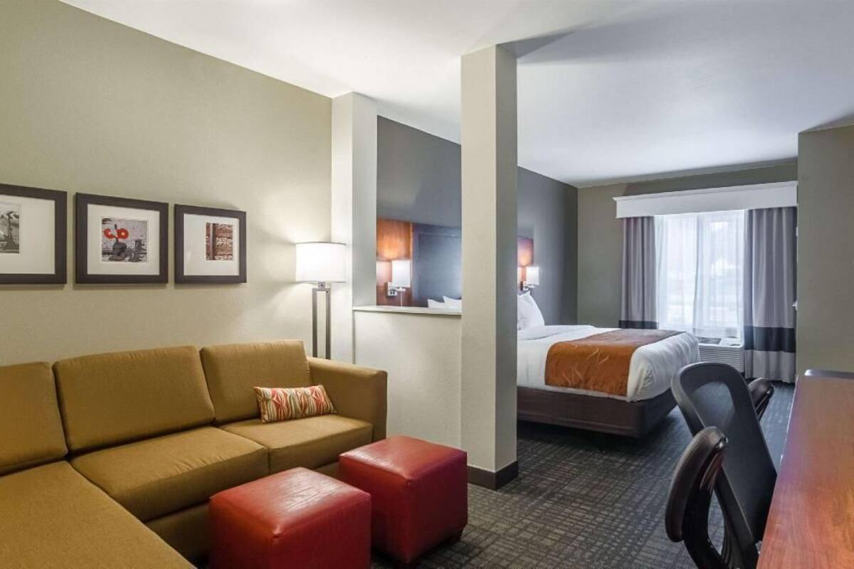 A hotel room with a sofa, ottoman, desk, chair, bed, and artwork on the wall.