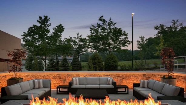 A modern outdoor seating area with a fire feature, stone wall, and trees in the background. - Embassy Suites by Hilton Berkeley Heights (Berkeley Heights)