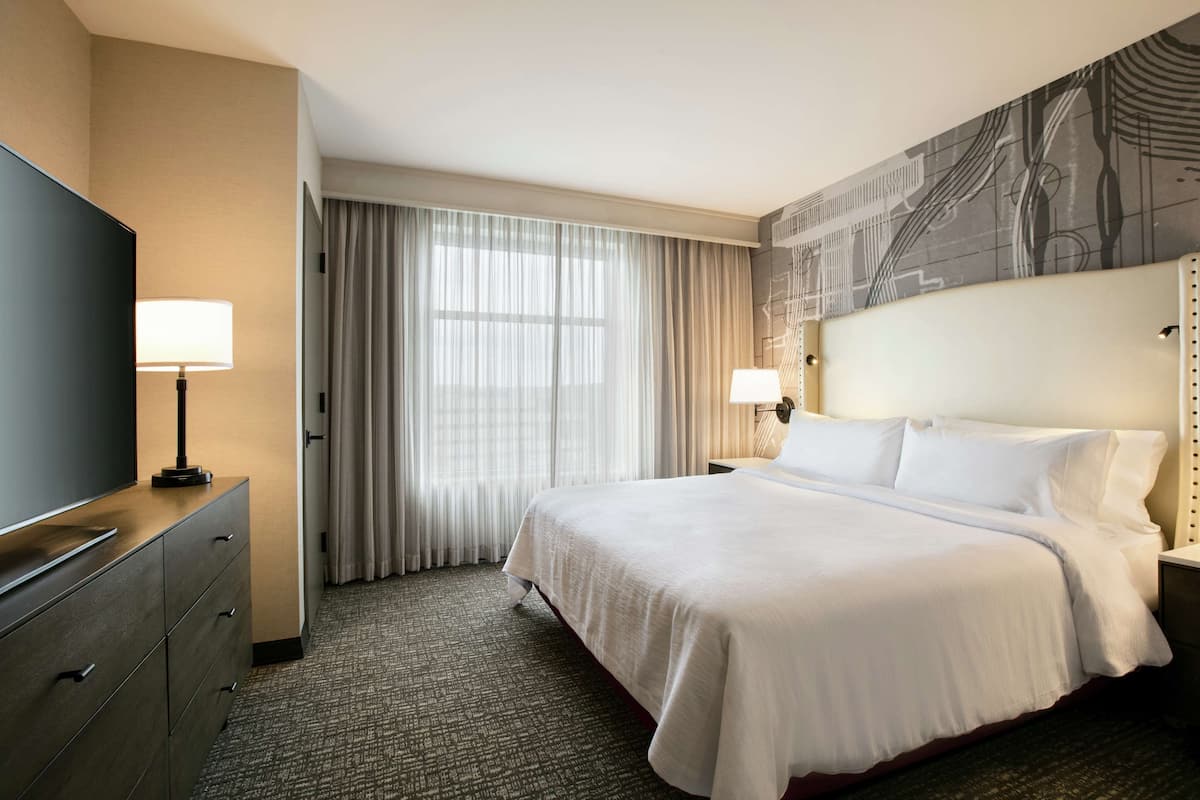 A hotel room with a large bed, a flat-screen TV, a desk with a lamp, and a view of the outdoors through the window.