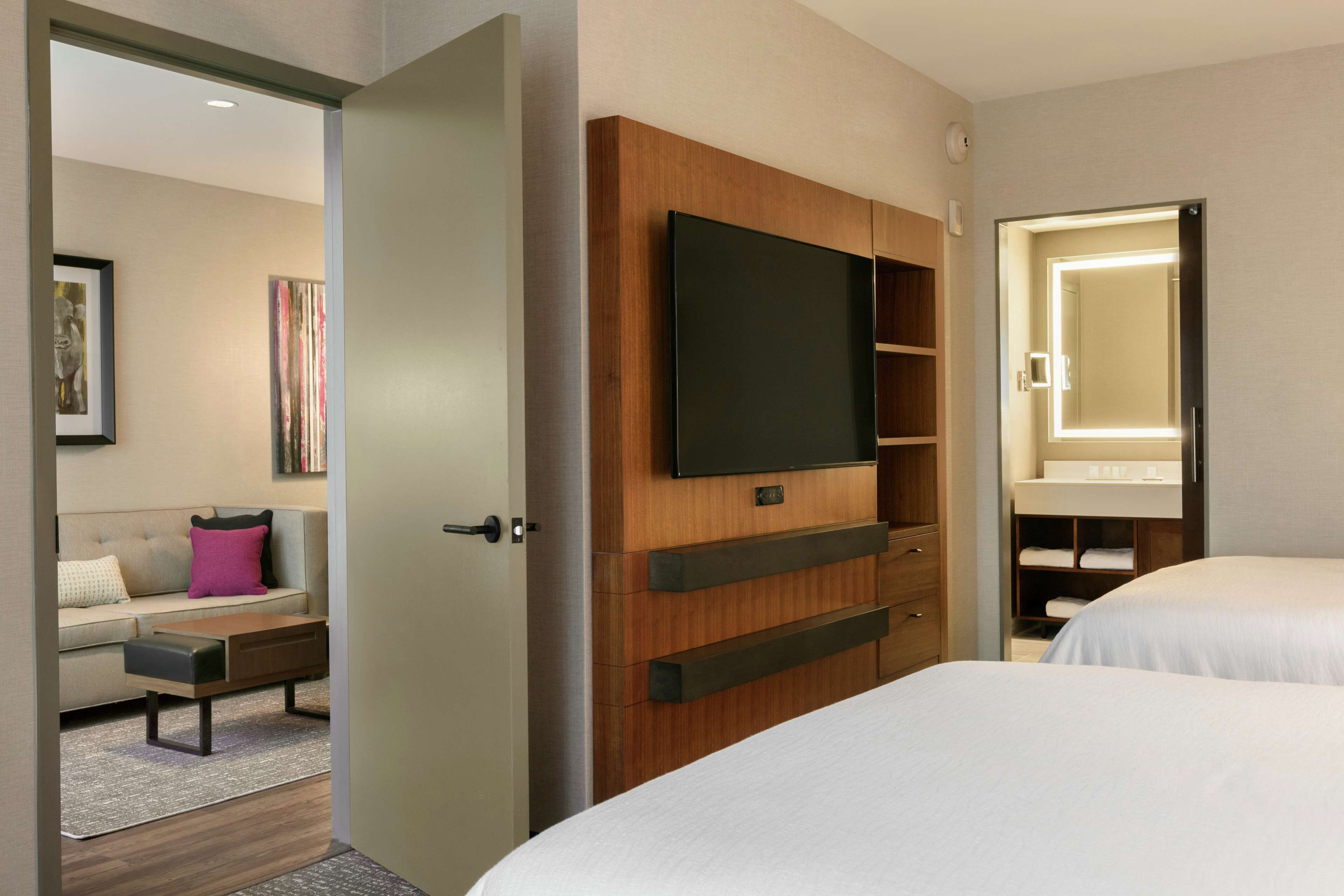 Ein Hotelzimmer mit einem Holzkoffer, einem Flachbildfernseher, einem Bett, einem Sofa und einem Badezimmer mit Spiegel.