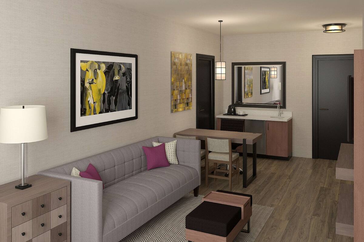 A modern hotel room with a grey sofa, a small desk, a mirror, and a wooden floor.