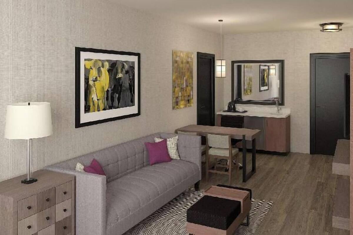A modern hotel room with a grey sofa, a small table, a desk with a mirror, a lamp, and artwork on the walls.