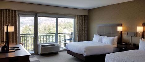 Een hotelkamer met twee bedden, een bureau, een stoel en een balkon met uitzicht op bomen.