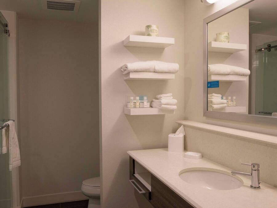 Un baño con cabina de ducha de vidrio, lavabo con espejo y repisas con toallas y artículos de aseo.