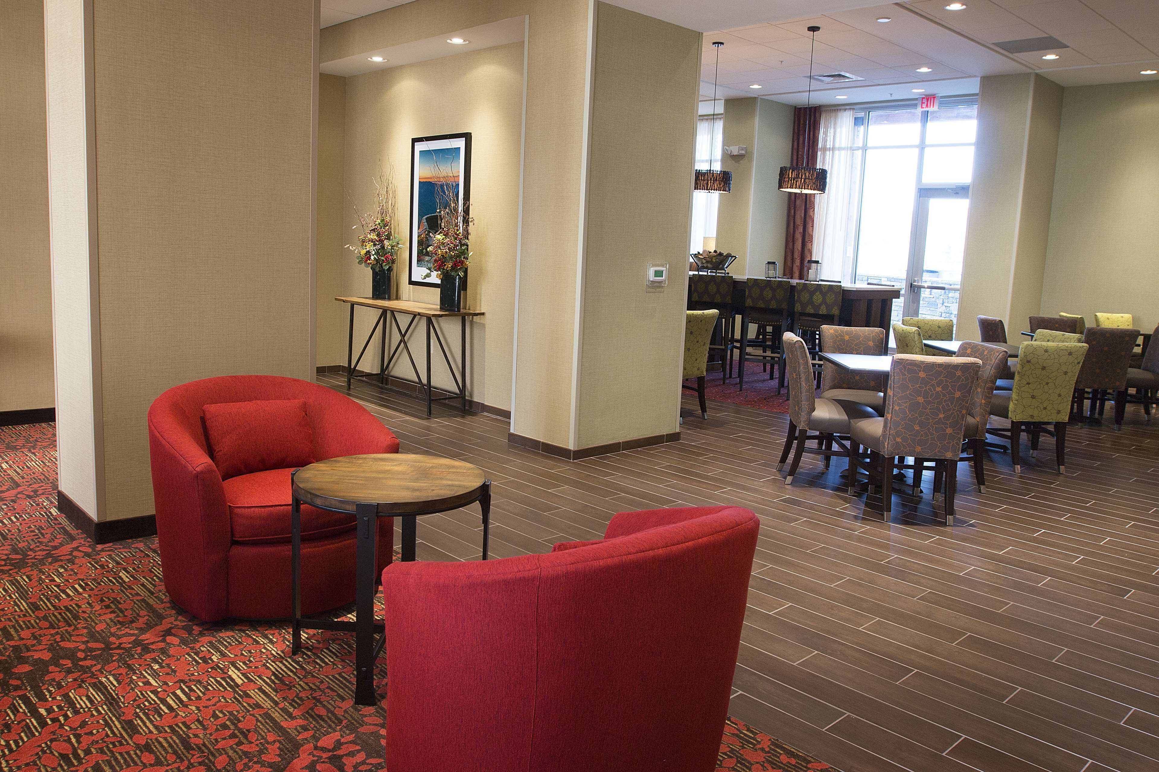 A hotel lobby with red armchairs, a coffee table, a dining area with tables and chairs, and a large window providing natural light.