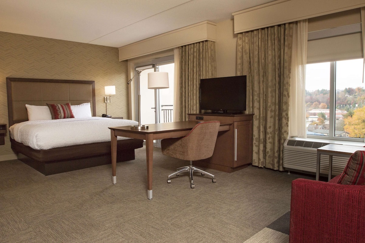 A hotel room with a bed, desk, chair, TV, and a view of the outside.