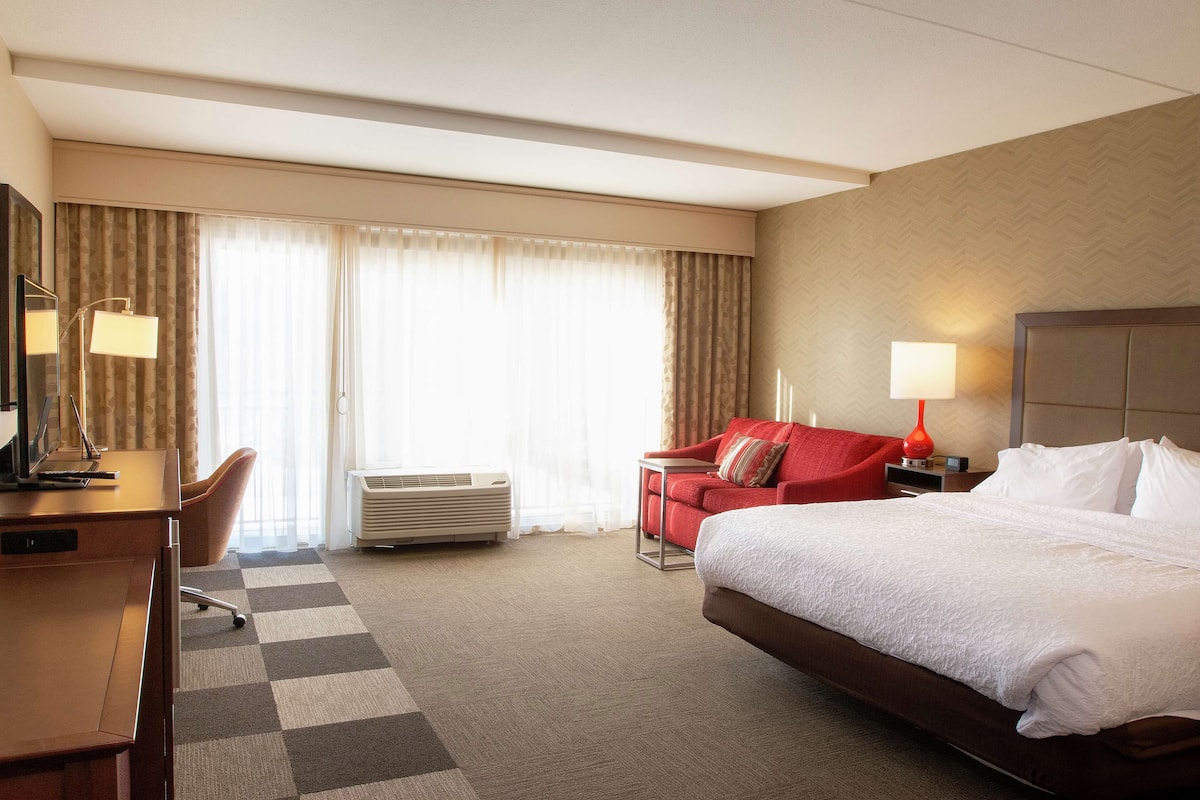 A hotel room with a large bed, a desk, a chair, a red sofa, and a window with curtains.