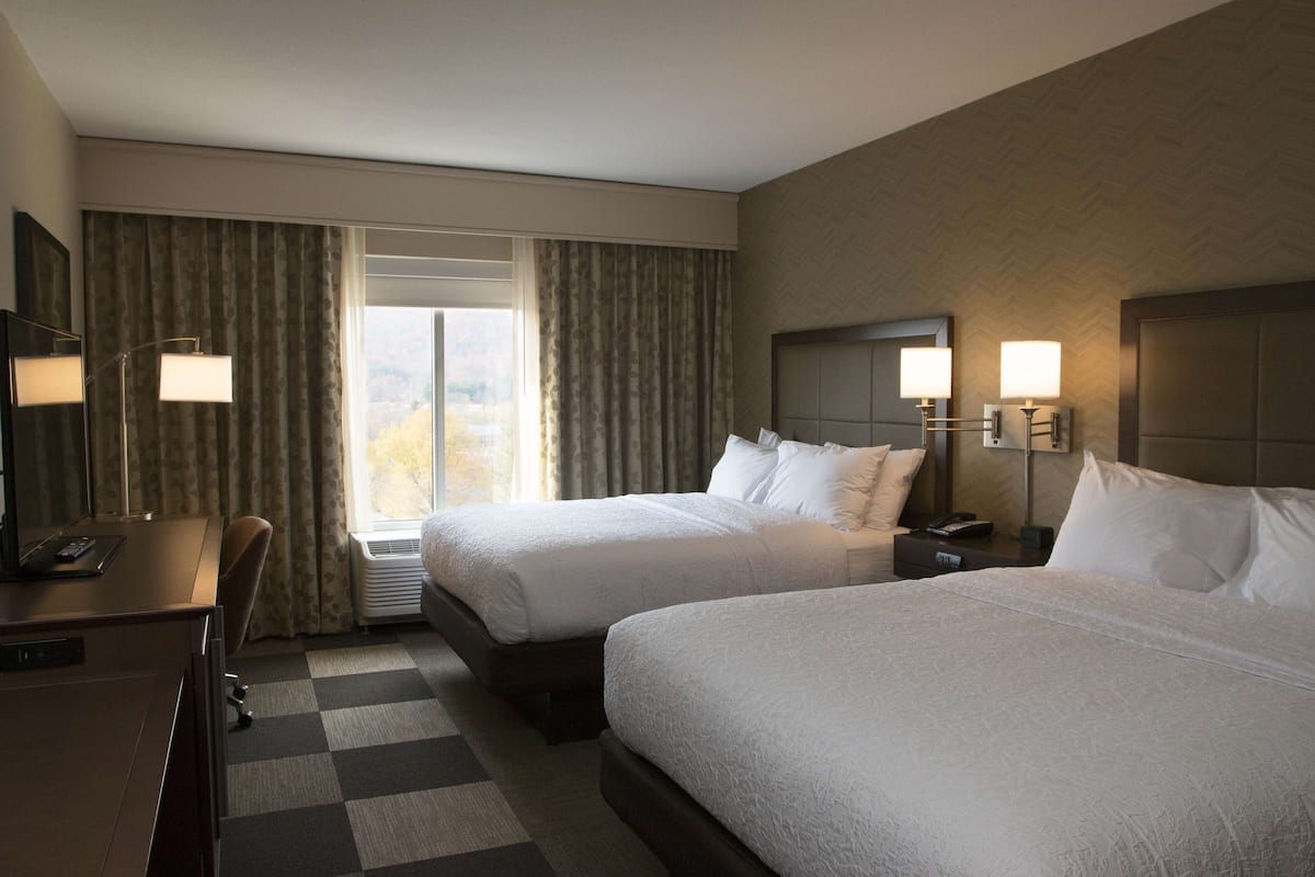 A hotel room with two beds, a desk, and a large window with curtains.