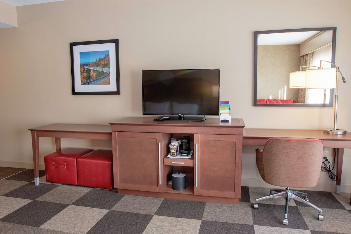 A hotel room with a large flat-screen TV, a desk with a lamp, a red chair, a coffee maker, and a framed picture on the wall.