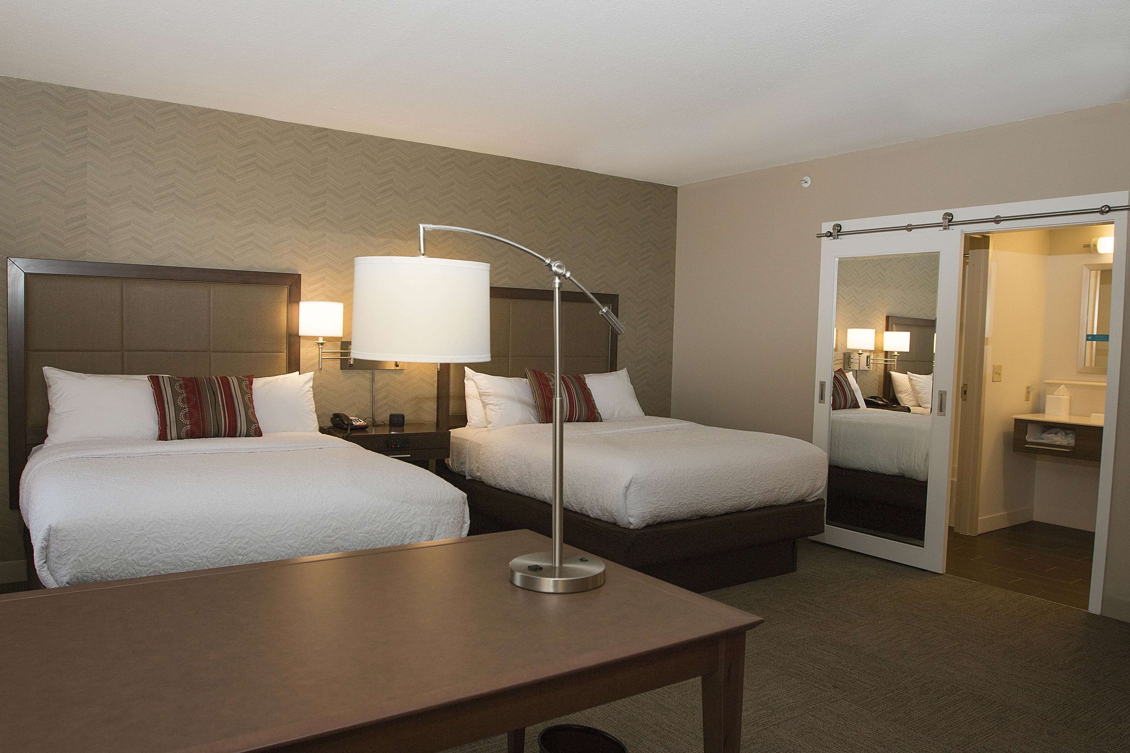 A hotel room with two beds, a desk, a lamp, and a mirror.
