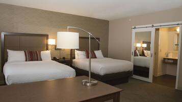 A hotel room with two beds, a desk, a lamp, and a mirror.