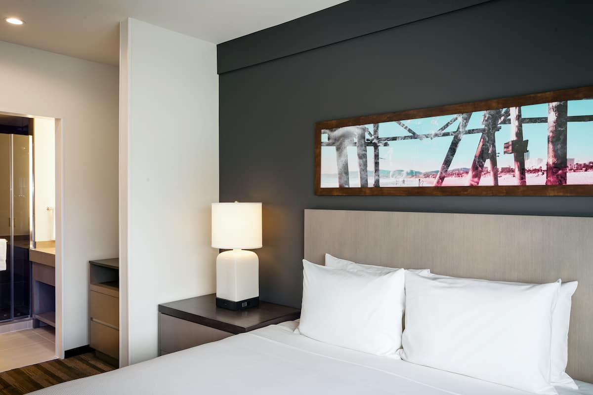 Una camera d'albergo con un letto, un comodino con una lampada, un quadro appeso al muro e una porta sul lato sinistro.