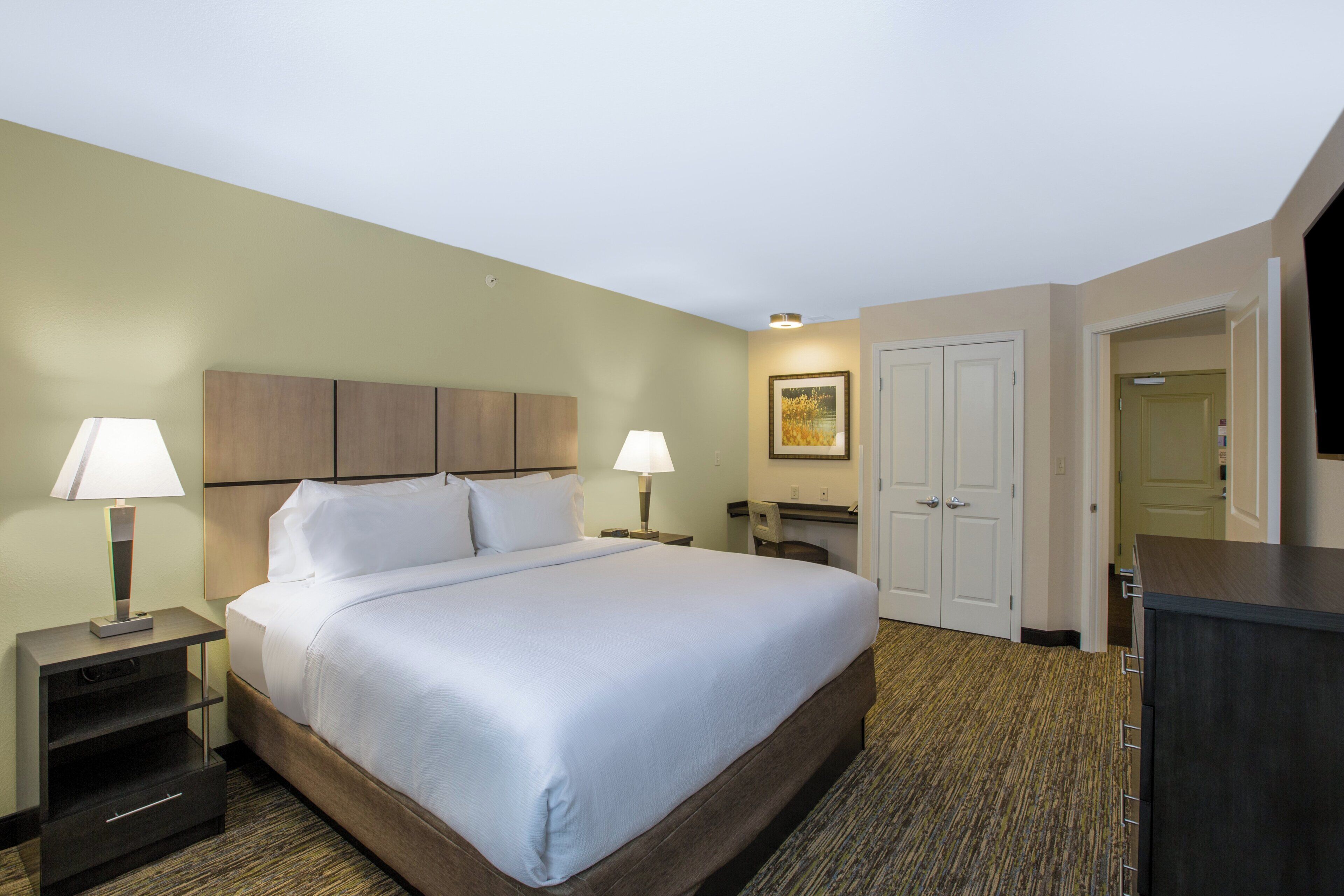 Quarto de hotel com uma cama grande, duas mesas de cabeceira com luminárias, uma escrivaninha, uma cadeira, um guarda-roupa e um quadro na parede.
