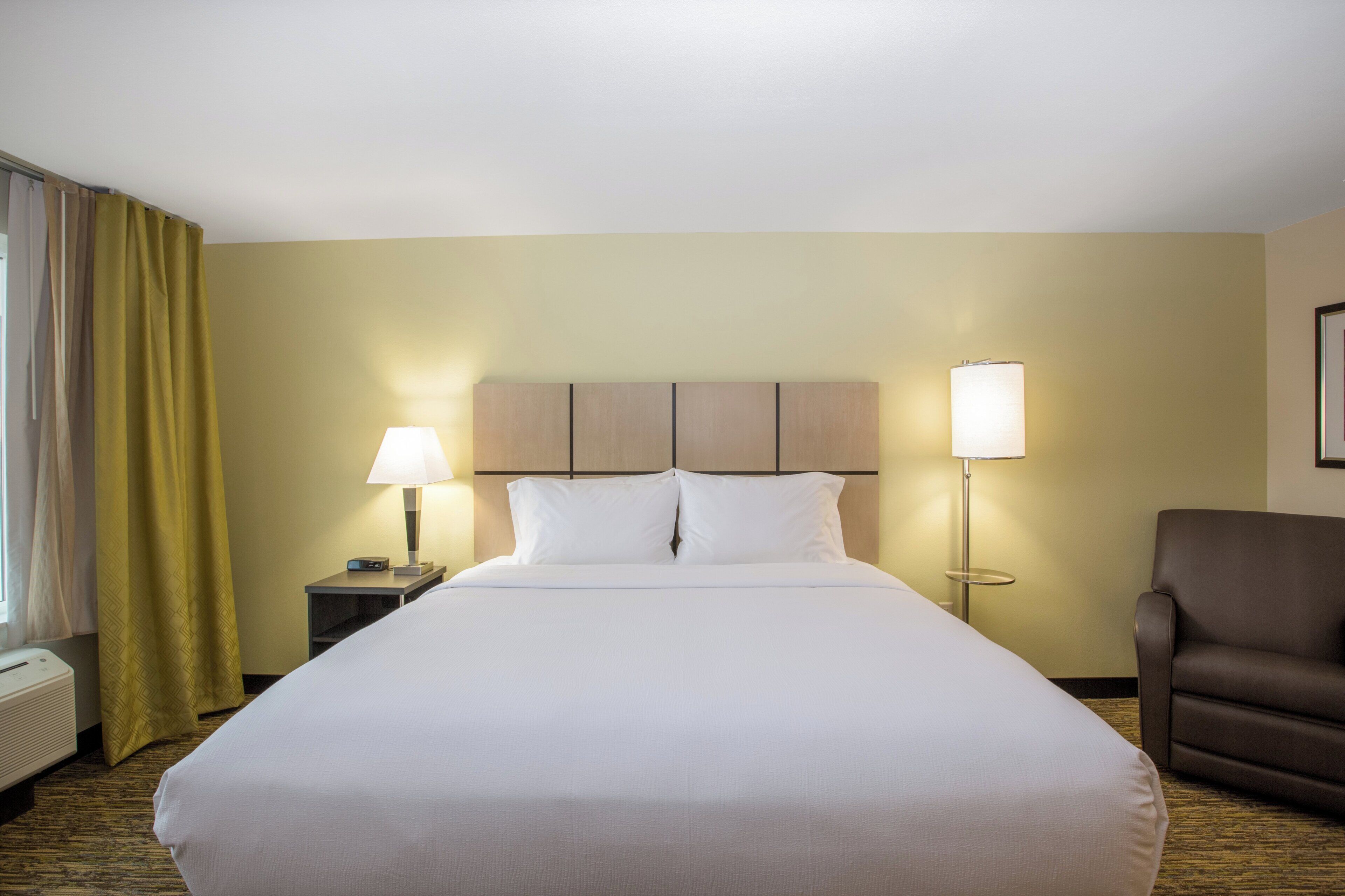 Une chambre d’hôtel avec un grand lit, deux lampes de chevet, une chaise et une fenêtre avec des rideaux.